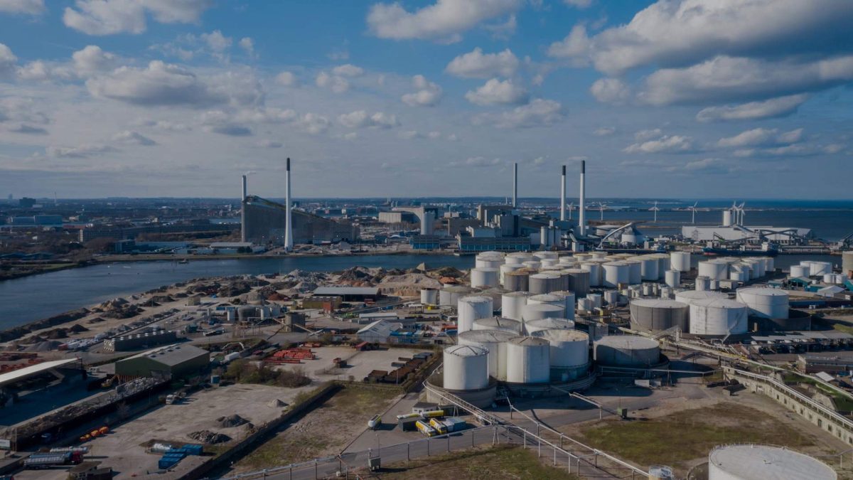 From an aerial perspective, the industrial area reveals numerous large white storage tanks and factories with tall chimneys along a river. The scene, ideal for asset inspection, unfolds beneath a partly cloudy sky with urban infrastructure in the distance.