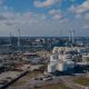 From an aerial perspective, the industrial area reveals numerous large white storage tanks and factories with tall chimneys along a river. The scene, ideal for asset inspection, unfolds beneath a partly cloudy sky with urban infrastructure in the distance.