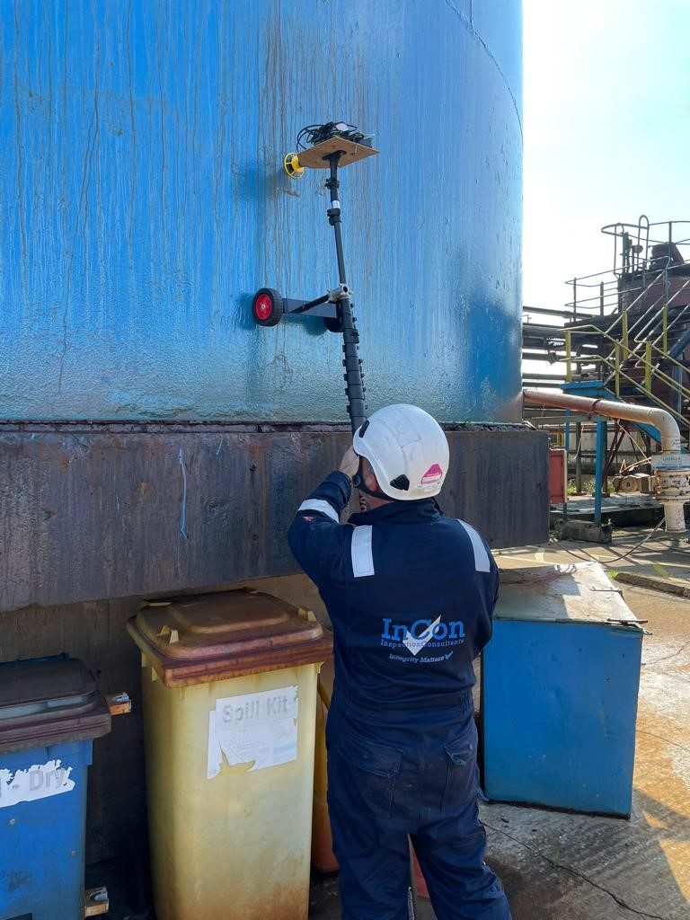 A person in protective gear and a hard hat carries out an inspection of a large blue industrial tank using a device mounted on a pole. Nearby, yellow containers labelled Spill Kit are visible, while the navy jacket with Incon on the back identifies the crew member.