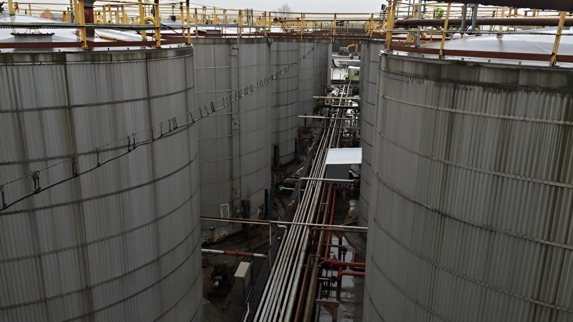 Large industrial storage tanks are arranged in rows at a facility. Metal pipes and walkways, maintained through rigorous condition monitoring, run between the tanks. Railings are visible on the top edges. The sky is overcast, indicating a cloudy day.