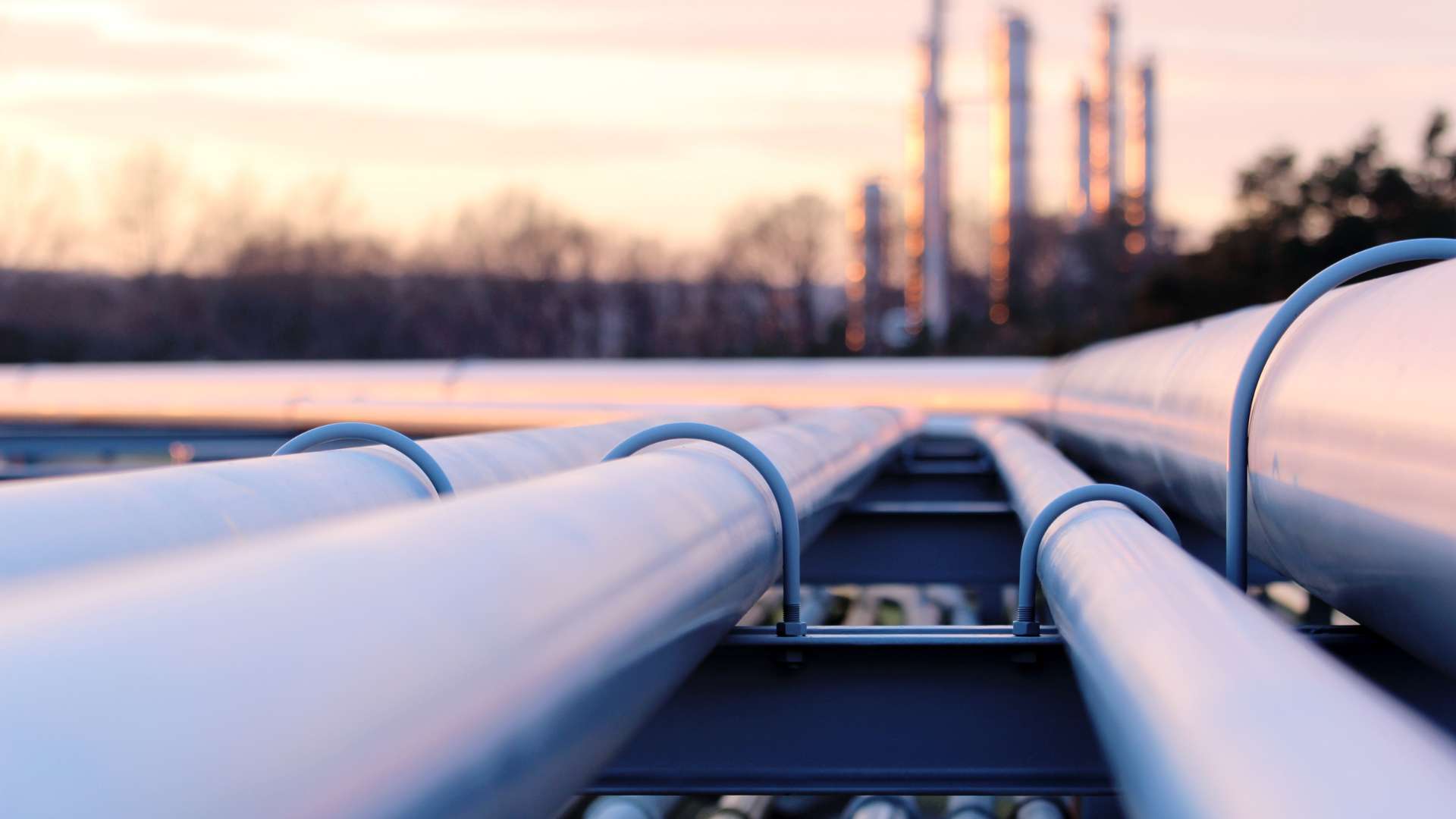 Close-up of large metal pipelines running parallel outdoors at sunset, ideal for asset inspection. The pipes, supported by brackets, extend towards an industrial facility with tall structures in the background. Trees create a blurred backdrop.