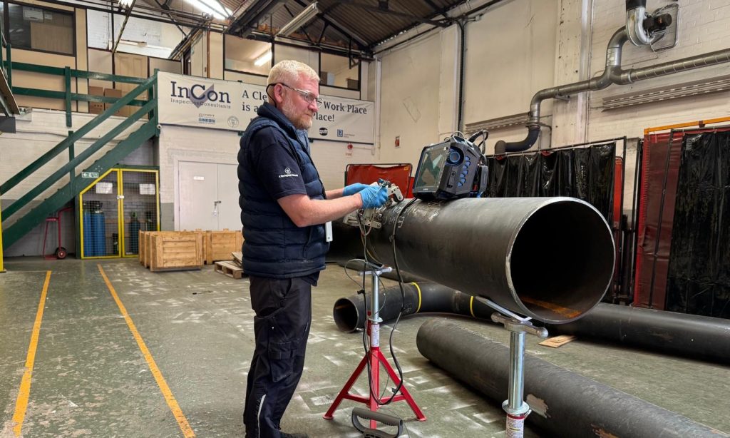 A man wearing safety glasses and gloves carries out a manual phased array inspection on a large metal pipe inside an industrial workshop, with equipment and safety signs visible in the background.
