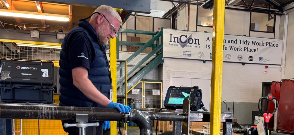 A man wearing spectacles, blue gloves, and a black waistcoat carries out a manual phased array inspection on a metal pipe using electronic equipment in an industrial setting. A sign reads, “A Neat and Tidy Workplace is a Safe Workplace!”.