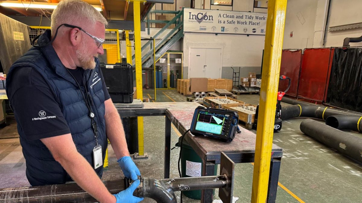 A man wearing safety glasses, gloves, and a vest carries out a manual phased array inspection using an ultrasonic testing device on a metal pipe in an industrial workshop. Various equipment and materials are visible in the background.