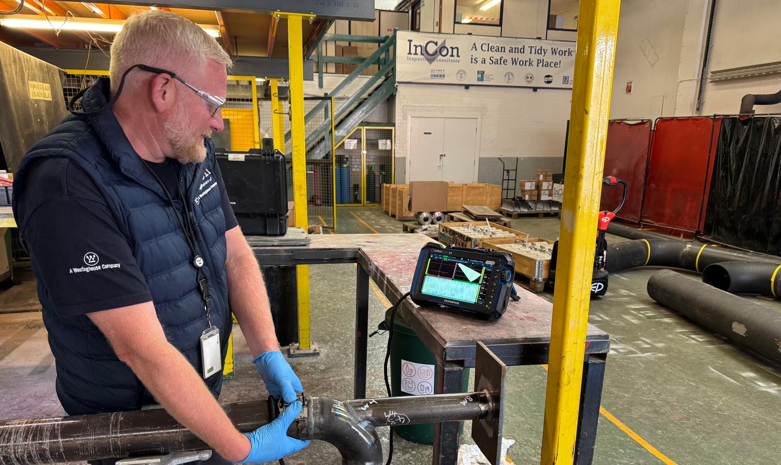 A man wearing safety glasses, gloves, and a vest carries out a manual phased array inspection using an ultrasonic testing device on a metal pipe in an industrial workshop. Various equipment and materials are visible in the background.